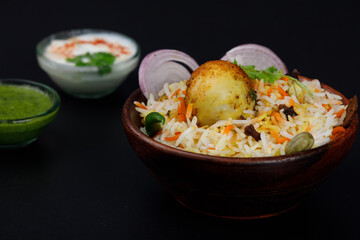 Egg biryani with fried egg served in a wooden bowl with Raita or curd or yogurt and green coriander chutney isolated on a black background