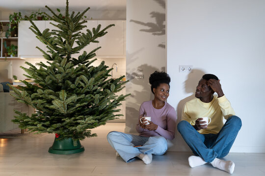 Happy african american couple resting after installing natural pine at home sit on floor, drink hot tea or cocoa and talk. Happy apartment owners prepare to celebrate christmas and new year together