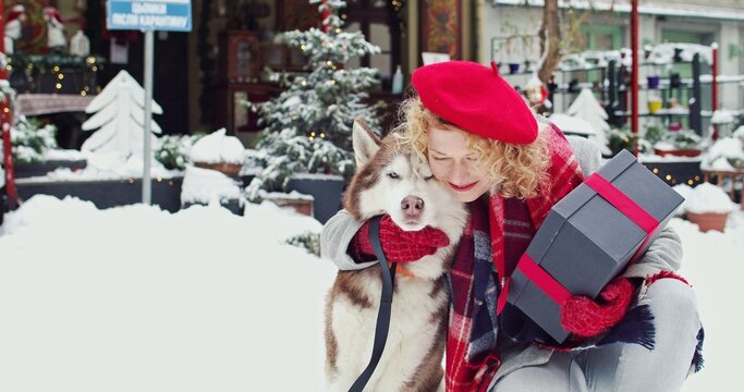 Beautiful young curly woman hugging a dog on decorated street, smiling and holding Christmas gifts. Christmas mood concept.