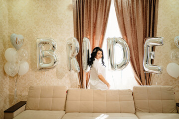 bride in white coat in a room of her house by sofa with balloons.