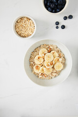 Bowl of porridge topped with banana and almonds,  a bowl of oats and a  bowl of blueberries on white marble countertop