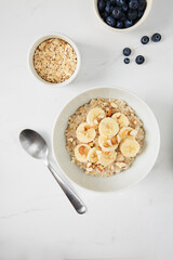 Bowl of porridge topped with banana and almonds,  a bowl of oats and a  bowl of blueberries on white marble countertop