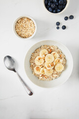 Bowl of porridge topped with banana and almonds,  a bowl of oats and a  bowl of blueberries on white marble countertop