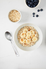Bowl of porridge topped with banana and almonds,  a bowl of oats and a  bowl of blueberries on white marble countertop