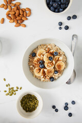 Bowl of porridge topped with banana, blueberries, almonds and cinnamon,  a bowl of pumpkin seeds and a  bowl of blueberries on white marble countertop