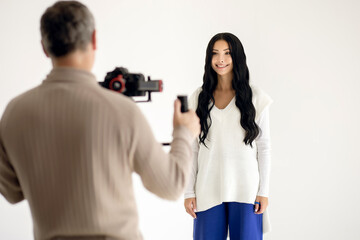 A girl blogger shoots a video blog in the studio on a white background with the help of a videographer with a professional video camera. Modern work as a blogger or journalist