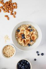 Bowl of porridge topped with banana, blueberries, almonds and cinnamon, a bowl full of oats and berries along with almonds and blueberries scattered on white marble countertop