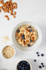 Bowl of porridge topped with banana, blueberries, almonds and cinnamon, a bowl full of oats and berries along with almonds and blueberries scattered on white marble countertop