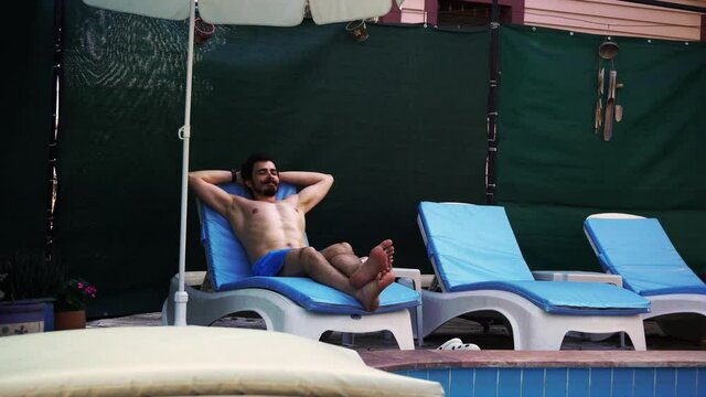 Relaxed man is lying on the sunbed next to the swimming pool and sunbathing with a smile on his face. A caucasian male person is resting in a hotel during his vacations. He is tanning with closed eyes