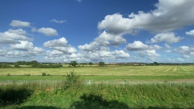 Wide shot countryside & farmland clouds and blue sky out of a window of train, tracking along flat british UK southern England  - stock footage video