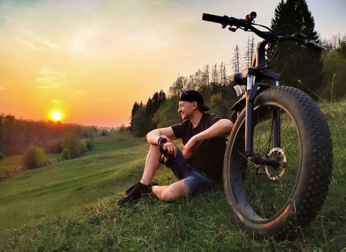 A man is sitting on the grass near a bicycle with thick wheels. Against the background of sunset.