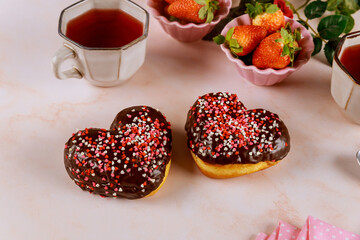 Two heart shaped donuts with white glaze and pink, red sprinkles with strawberry and cup of tea.