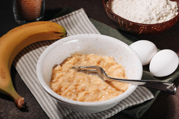 Banana puree in ceramic bowl and ingredients for cooking banana bread on brown background