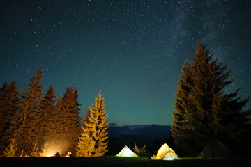 Silhouette of lonely hiker resting besides burning bonfire near illuminated tourist tents on camping site in dark mountains under night sky with stars. Active lifestyle and outdoor living concept