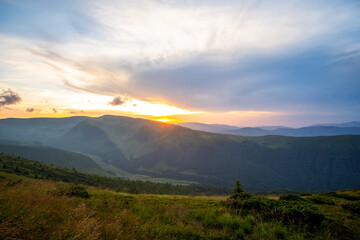 Obraz premium Summer evening mountain landscape with grassy hills and distant peaks at colorful sunset.