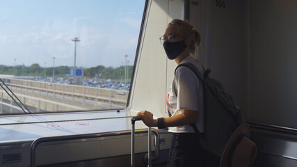 Pretty woman traveler wearing face mask on the way to airport terminal, standing and looking into the window. Female passenger traveling by train. Transportation and new normal travel concept.