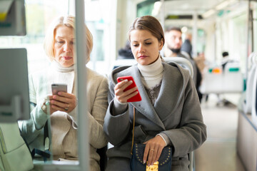 Two women passengers of different ages using mobile phones while traveling by public transport in sunny autumn day..