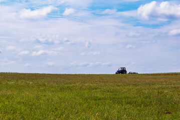 Naklejka premium Tractor working in the field, plowing the land, preparing for sowing. View of the field and tractor in the distance against a blue sky with clouds