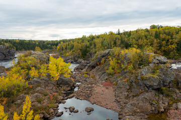 Rocky river with fast moving water. The river banks are covered in autumn foliage, large boulders, and tall trees. The sky is blue with lots of clouds. The leaves are red, yellow, and orange in color.