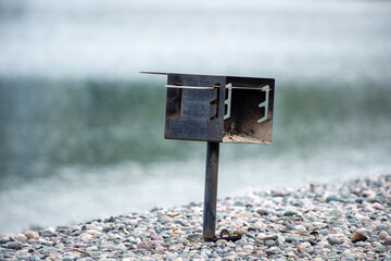 A metal barbeque grate on a single pedestal located on a pebbly beach. The ocean is in the background with waves rolling in the cove. The metal grate is empty of coal and firewood.  - Powered by Adobe