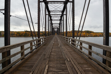 Steel trestle bridge with wooden deck over a large river in Newfoundland.  The bridge is for foot traffic and ATV usage. The sky is clear blue and land and houses can be seen in the background. 
