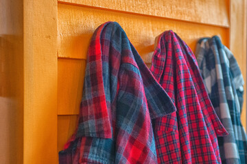 Three plaid shirts hung on wooden pegs on the inside of a vibrant yellow wooden country shed made of clapboard. The men's shirts are green, blue, and red in color with long sleeves and collars. 