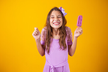 Happy fair-skinned child girl eats purple ice cream flavored with grapes, clenches her fist with...