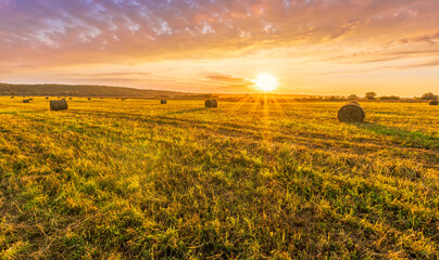 Obraz premium Scenic view at picturesque burning sunset in a green shiny field with hay stacks, bright cloudy sky , trees and golden sun rays, summer valley landscape