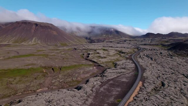 Turning aerial shot over dramatic Icelandic landscape: old lava fields. Mountains and house in distance with road winding below.