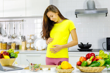 Caucasian young pregnant painful female mother in casual outfit standing holding tummy belly having stomachache while learning to cook vegetable salad online from laptop computer in kitchen at home