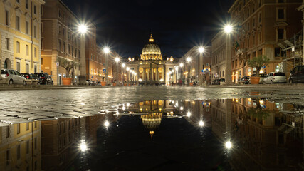 Vista nocturna de la cúpula de la Basílica de San Pedro en El Vaticano, reflejada en un charco de la Vía della Conciliazione.