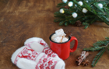 Red mug with hot chocolate with marshmallow and mittens nearly and branches of christmas tree on wooden table