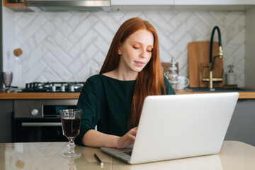 Pretty female freelancer working typing on laptop computer sitting at table with glass of alcoholic...