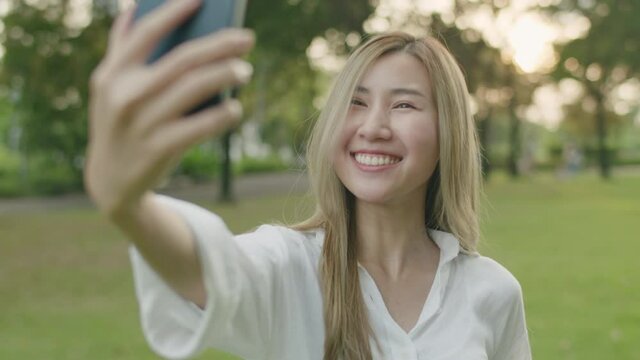 Smiling Asian woman taking selfie photo on smartphone at a public park during sunset, Happy woman influencer using mobile phone camera take pictures.