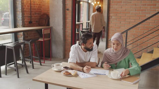 High-angle of hazel-eyed Caucasian woman wearing hijab and bearded Biracial man sitting at table in busy modern cafe, doing paperwork, people walking on background