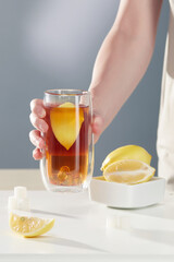 Shot of a female hand holding a glass of black tea with lemon. There are three cubes of refined sugar, a slice of lemon and lemons on the table. Items on the gray background.