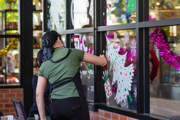 Asian women decorate beautiful stickers on window glass in front of cafe to welcome Christmas and New Year. Prepare to decorate the storefront to be beautiful during the important holidays of year.