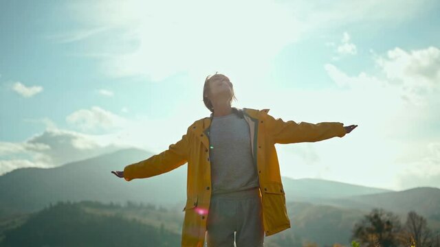 slow motion portrait Young woman wearing a yellow jacket raising her arms into blue sky and enjoying view above clouds on top of mountain.