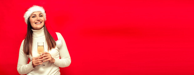 Fototapeta premium Portrait of a young smiling woman in Santa's cap and a glass of champagne in her hands on a red background