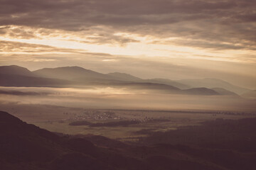 Vintage color landscape with low clouds above village in mountain valley among mountains silhouettes under dawn cloudy sky. Atmospheric scenery of countryside in low clouds in sepia and faded tones.