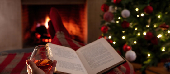 Christmas relaxation at home. Man with a book and a brandy glass, burning fireplace background.