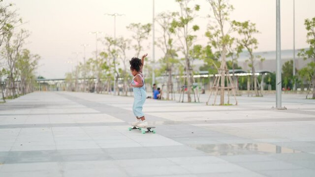 Cheerful little African child girl kid skateboarding at park in the city. Happy cute preschool girl enjoy and having fun outdoor lifestyle practicing extreme sport longboard skating on summer vacation