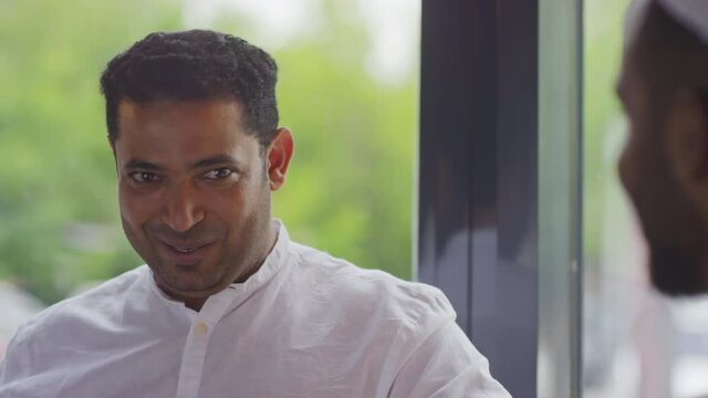 Shoulder-up of young Middle Eastern man wearing white shirt sitting in cafe by window at daytime, talking to cropped friend, smiling