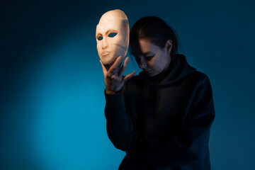 Hiding behind a mask, a young woman in a dark hoodie hides her face with a mask, self-identification problems and impostor syndrome. Portrait in the studio on a dark gray background. - obrazy, fototapety, plakaty