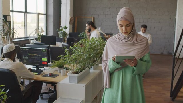 Tracking medium of pensive Caucasian woman wearing green dress and hijab, using tablet computer on-the-go, busy people walking around office at daytime