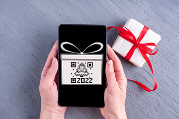 Hands holding a mobile phone with a black screen with a drawn gift box and a qr code in the shape of a Christmas tree and a gift box with a red ribbon on a gray background. Christmas online shopping.