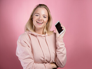 laughing young woman shows the phone screen and smiles . An attractive young woman in a pink hoodie uses a smartphone, a photo on pink.