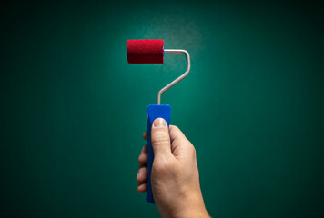 Man's hand holding red paint roller against green wall background, empty space for your text, banner, copyspace, advertisement or your design.