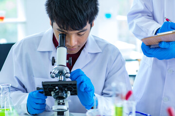 Asian young university male scientist student in white lab coat safety goggles and rubber gloves using microscope magnifying science sample in laboratory while teacher holding clipboard rating score