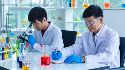 Fototapeta premium Portrait shot of Asian young male scientist in white lab coat and rubber gloves sitting smiling using microscope in laboratory while colleague wearing safety goggles writing result on paper clipboard